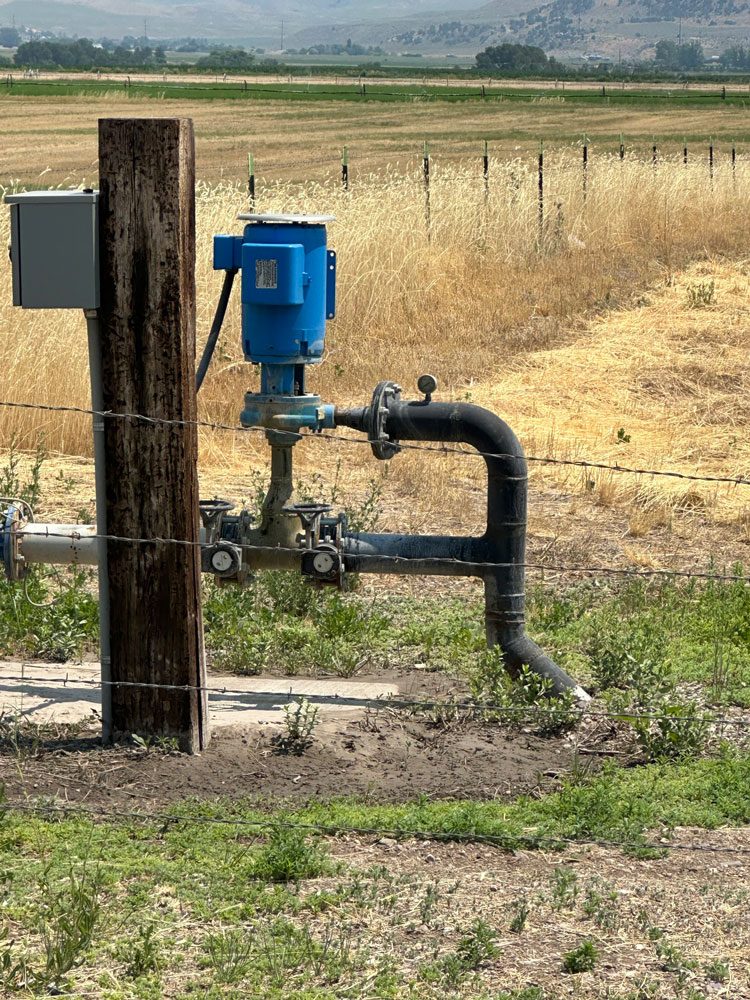 A blue booster pump is attached to a wooden utility pole, connected to a network of pipes, set against a backdrop of dry grass and farmland.
