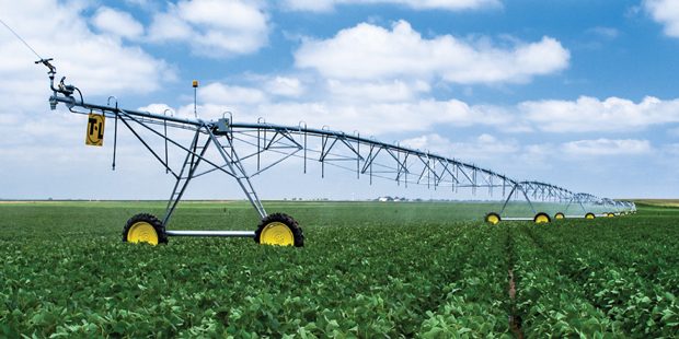 A large center pivot irrigation system spans across a green field, spraying water over crops under a clear blue sky.