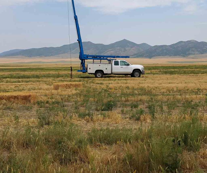 A white truck equipped with a blue drilling rig is parked in an open field, surrounded by natural scenery and distant mountains.