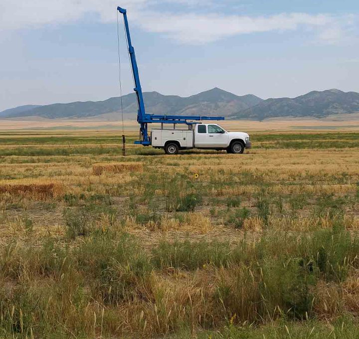 A white truck equipped with a blue drilling rig is parked in an open field, surrounded by natural scenery and distant mountains.