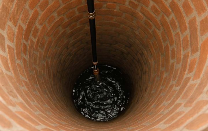 An interior view of a deep brick-lined well with a dark pipe extending downward, showcasing the circular structure and texture of the bricks.