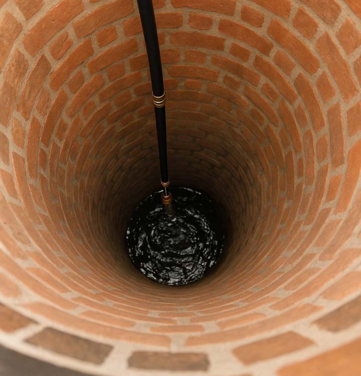 An interior view of a deep brick-lined well with a dark pipe extending downward, showcasing the circular structure and texture of the bricks.