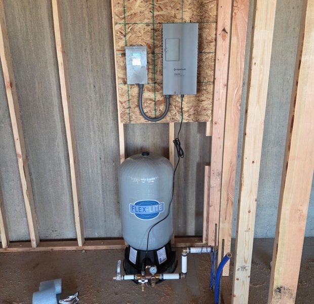 A new Flex-Lite well pressure tank and Pentair control boxes mounted on a wooden board inside an unfinished garage or utility room.