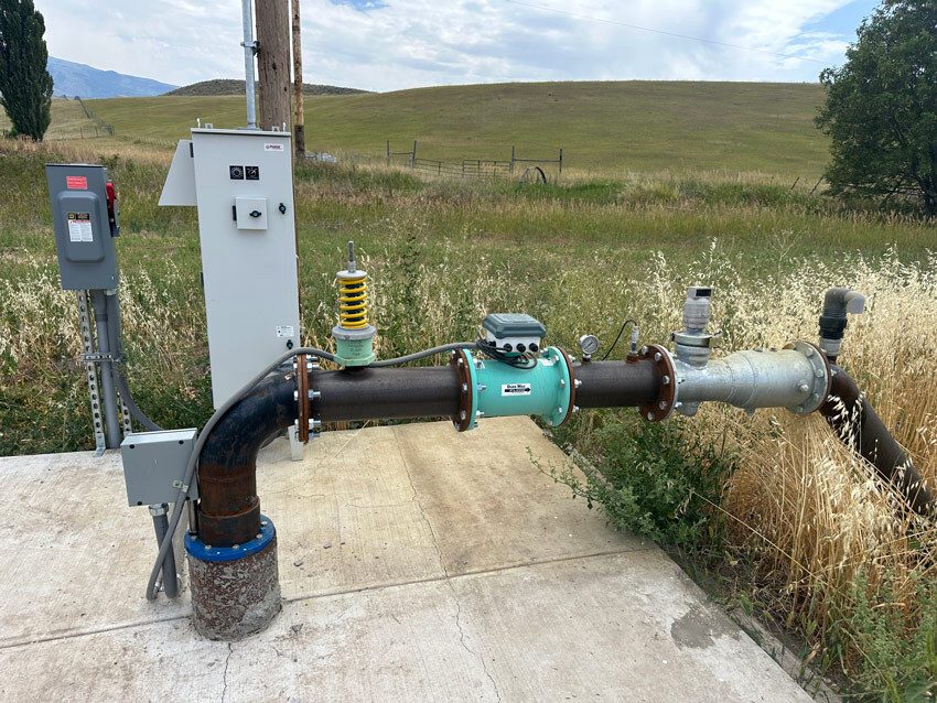 A robust water pump system with a control box and various pipes is installed on a concrete slab, surrounded by dry grass and open fields.
