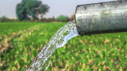 A close-up of water gushing out of a pipe, highlighting its flow into a lush green field with trees in the distance.