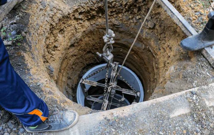 A deep well is being installed, with a cylindrical component suspended by ropes and chains, lowered into an excavated hole.