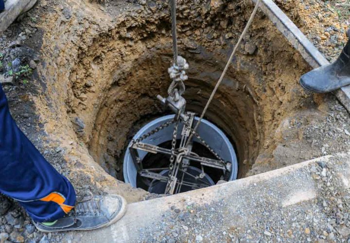 A deep well is being installed, with a cylindrical component suspended by ropes and chains, lowered into an excavated hole.
