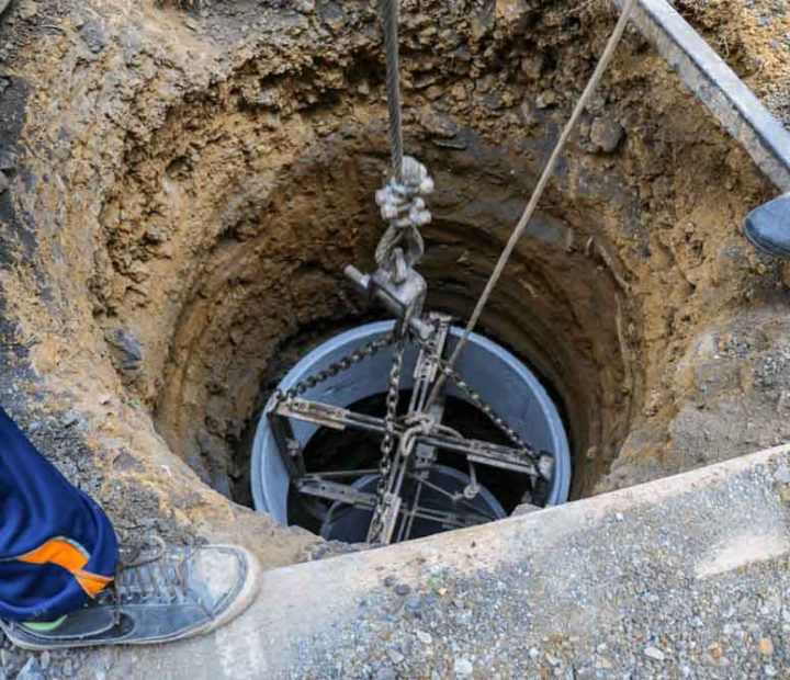 A deep well is being installed, with a cylindrical component suspended by ropes and chains, lowered into an excavated hole.