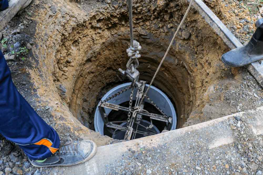 A deep well is being installed, with a cylindrical component suspended by ropes and chains, lowered into an excavated hole.