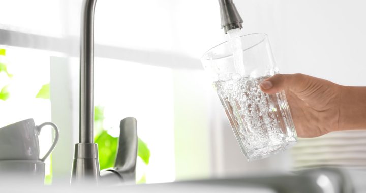 A person's hand holding a clear glass under a modern kitchen faucet, filling it with fresh, sparkling water.