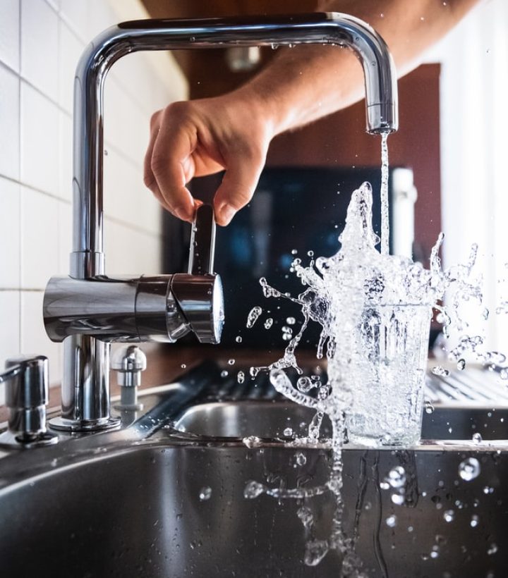 A person fills a glass with water from a modern kitchen faucet, capturing the dynamic motion of water splashing into the glass.