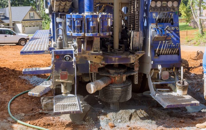 The rear of a blue drilling truck shows its powerful rotary head and auger actively drilling into the red clay soil of a residential construction site.