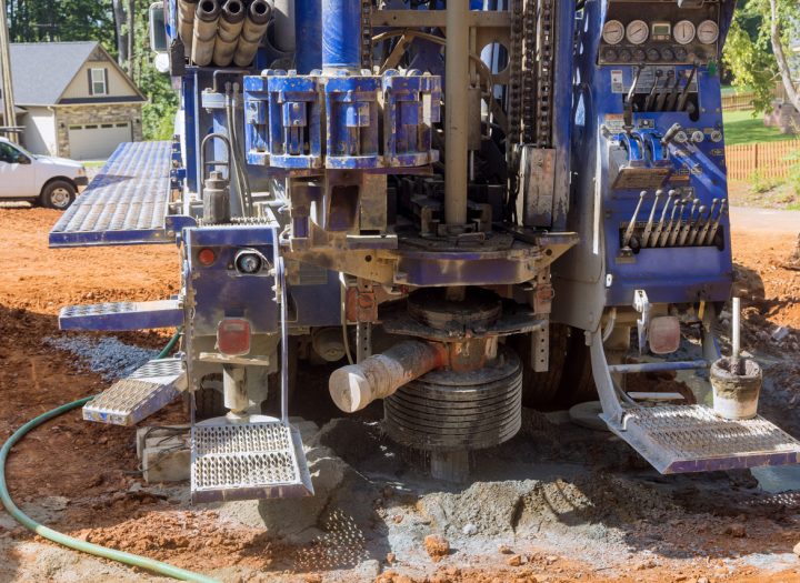The rear of a blue drilling truck shows its powerful rotary head and auger actively drilling into the red clay soil of a residential construction site.
