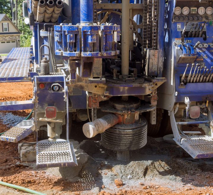 The rear of a blue drilling truck shows its powerful rotary head and auger actively drilling into the red clay soil of a residential construction site.