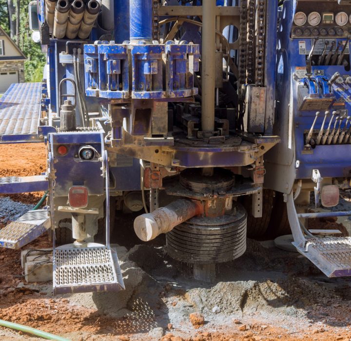 The rear of a blue drilling truck shows its powerful rotary head and auger actively drilling into the red clay soil of a residential construction site.
