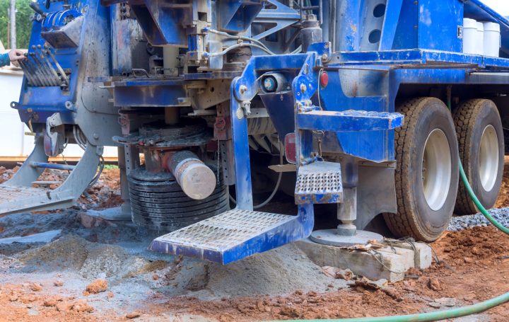The rear section of a large blue drilling truck, showing its outriggers extended onto wooden blocks for stability on a muddy work site.