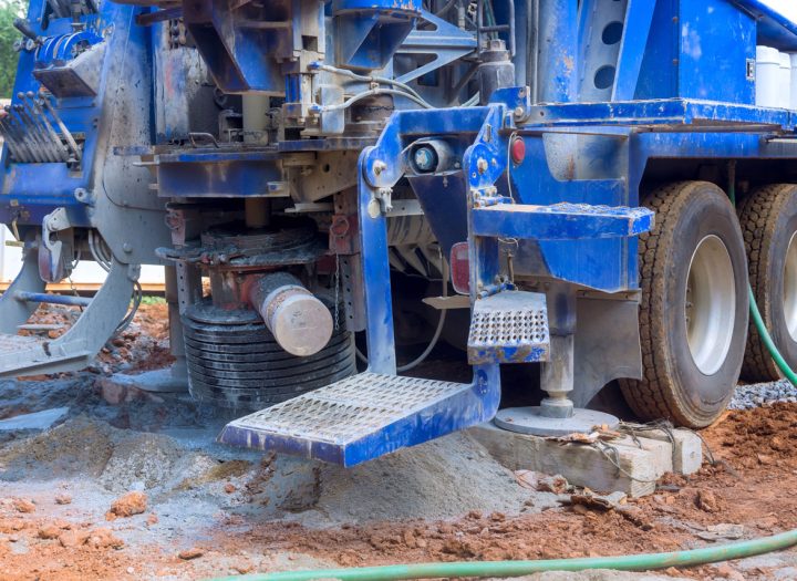The rear section of a large blue drilling truck, showing its outriggers extended onto wooden blocks for stability on a muddy work site.