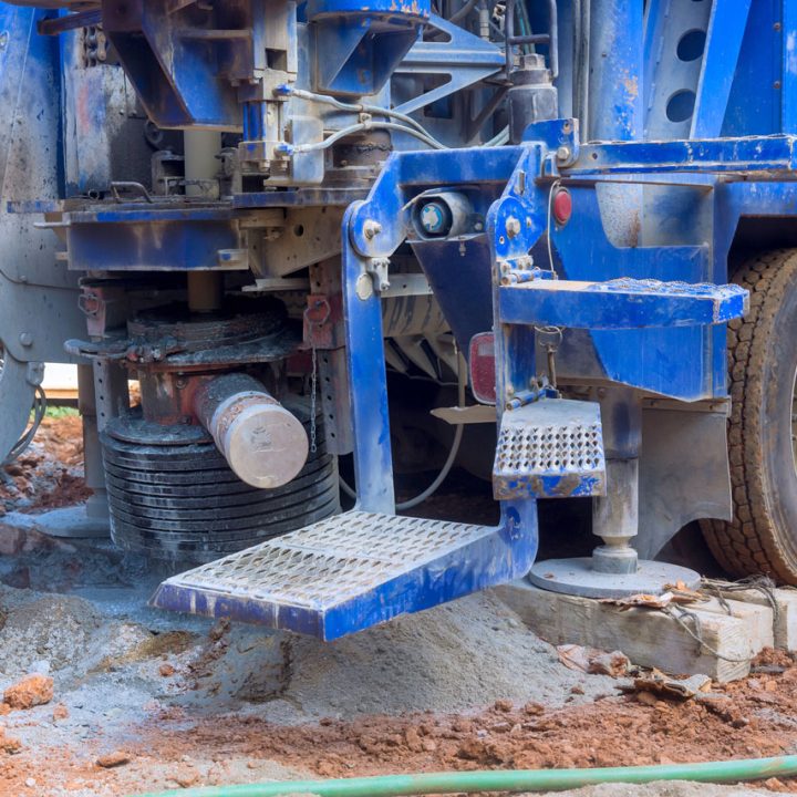The rear section of a large blue drilling truck, showing its outriggers extended onto wooden blocks for stability on a muddy work site.