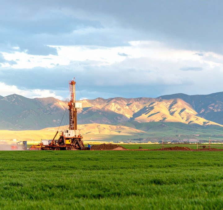 A drilling rig works in a vibrant green field, with sunlit mountains and a dramatic cloudy sky creating a scenic backdrop.