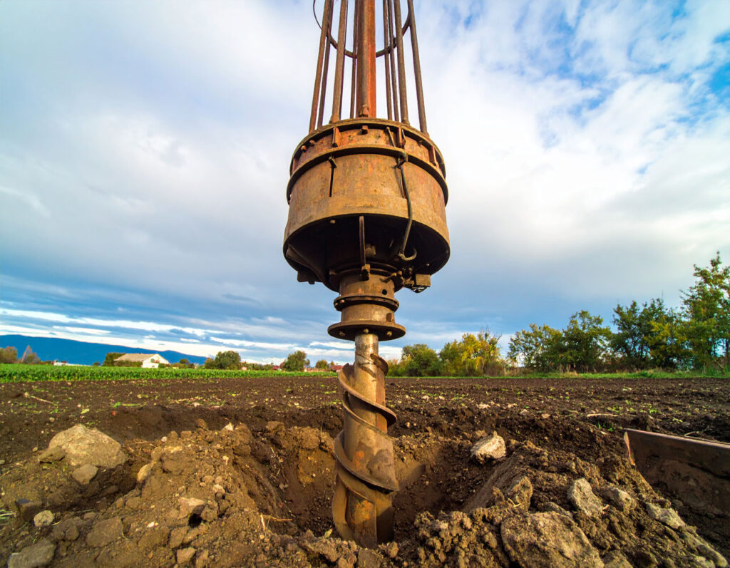 A low-angle, close-up shot of a heavy-duty auger drill bit poised over a freshly drilled hole in a plowed field, with a cloudy sky above.