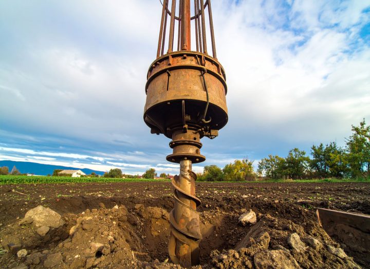A low-angle, close-up shot of a heavy-duty auger drill bit poised over a freshly drilled hole in a plowed field, with a cloudy sky above.