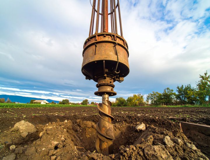 A low-angle, close-up shot of a heavy-duty auger drill bit poised over a freshly drilled hole in a plowed field, with a cloudy sky above.