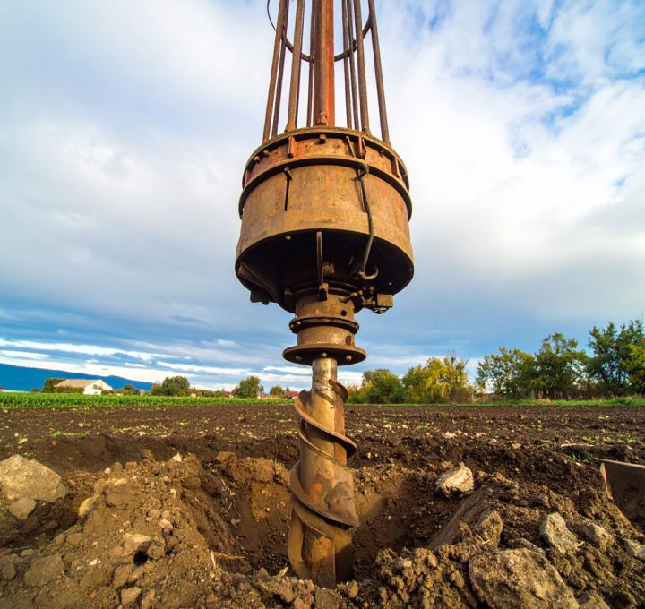 A low-angle, close-up shot of a heavy-duty auger drill bit poised over a freshly drilled hole in a plowed field, with a cloudy sky above.