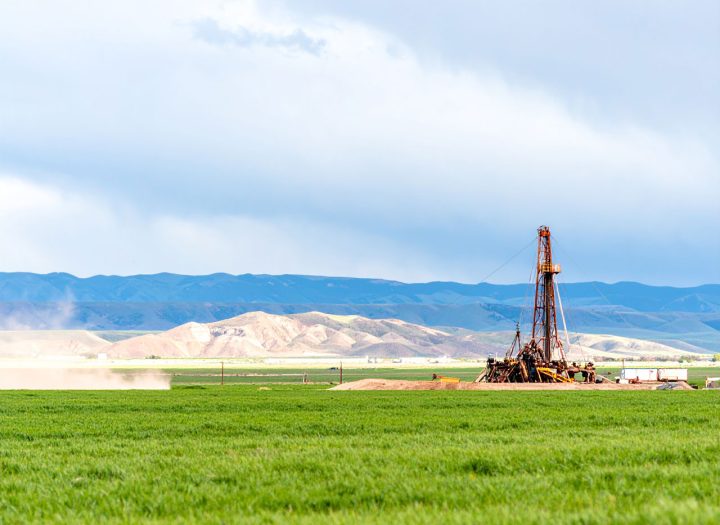 A wide shot of a drilling rig operating in a lush green field, with rolling hills and mountains under a cloudy sky in the background.