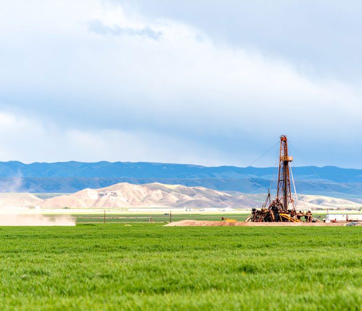 A wide shot of a drilling rig operating in a lush green field, with rolling hills and mountains under a cloudy sky in the background.