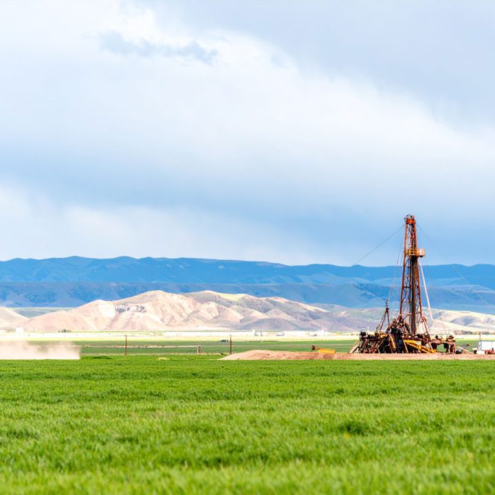 A wide shot of a drilling rig operating in a lush green field, with rolling hills and mountains under a cloudy sky in the background.