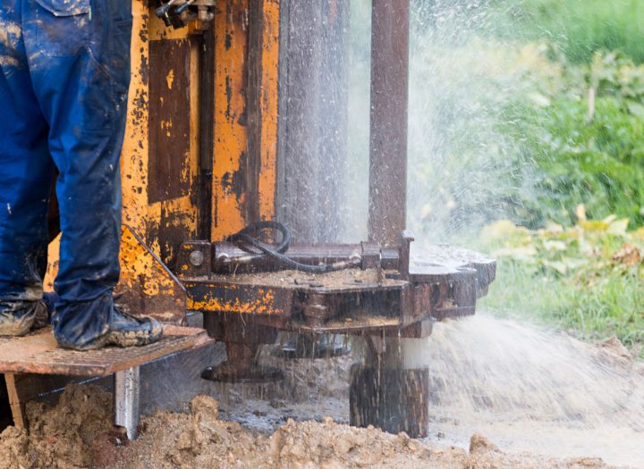 A close-up view of a worker's legs in muddy boots standing on a drilling platform as water sprays from the borehole during the drilling process.