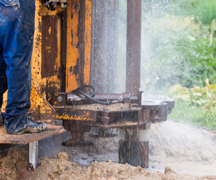 A close-up view of a worker's legs in muddy boots standing on a drilling platform as water sprays from the borehole during the drilling process.