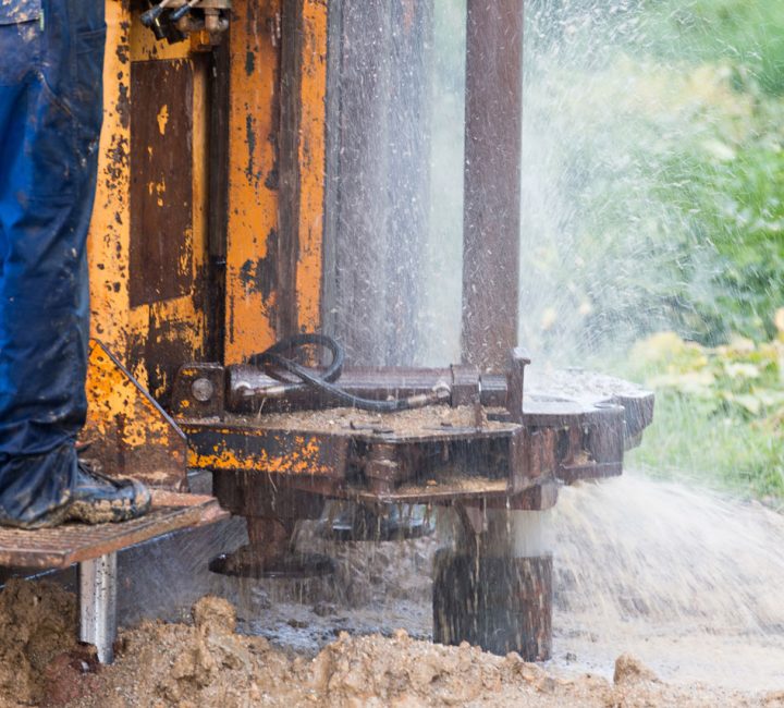 A close-up view of a worker's legs in muddy boots standing on a drilling platform as water sprays from the borehole during the drilling process.