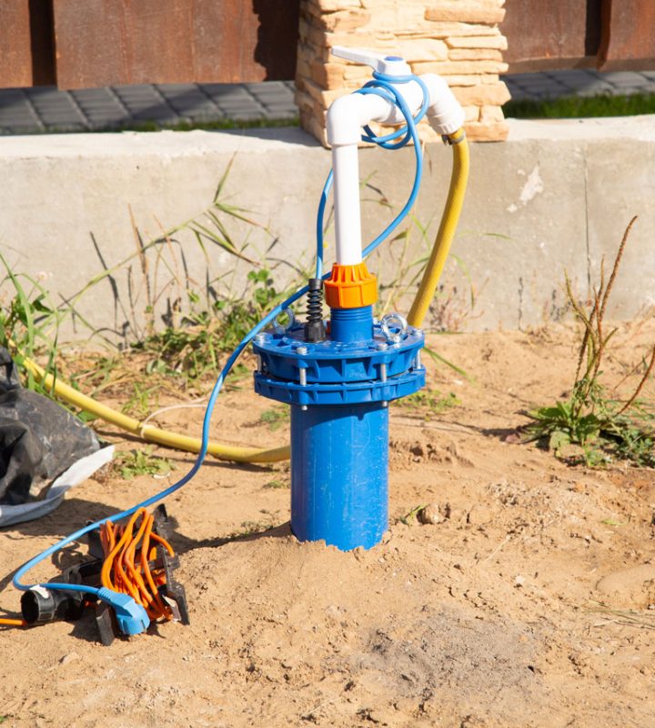 A portable blue well pump with white PVC piping and an orange extension cord sits on sandy ground near a building's foundation.