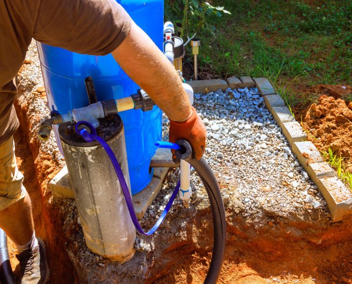 A worker wearing gloves lowers a submersible water pump into a well casing next to a large blue pressure tank during an installation.
