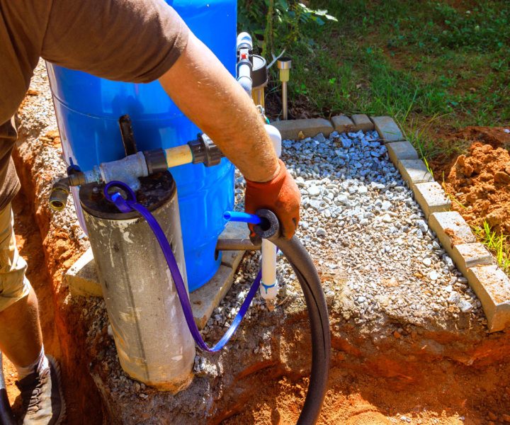 A worker wearing gloves lowers a submersible water pump into a well casing next to a large blue pressure tank during an installation.