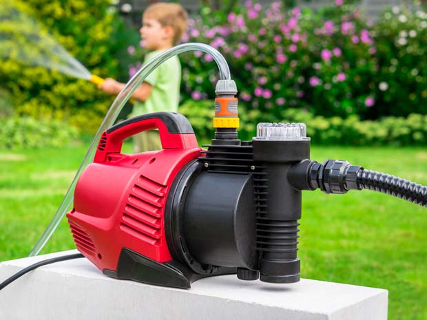 A red and black portable water pump is placed outdoors, connected to a hose, set against a backdrop of greenery and flowers.