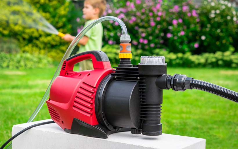 A red and black portable water pump is placed outdoors, connected to a hose, set against a backdrop of greenery and flowers.