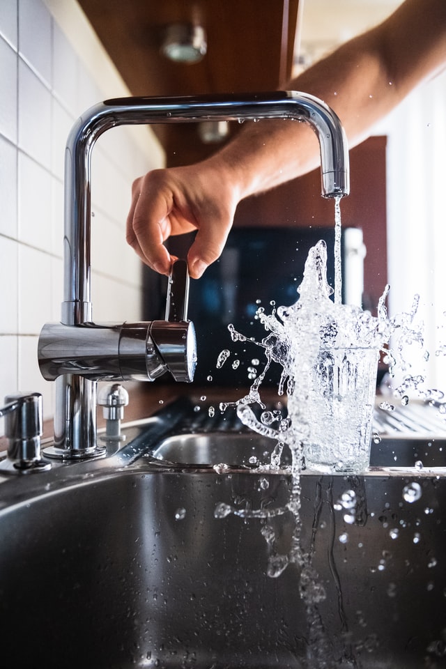 A person fills a glass with water from a modern kitchen faucet, capturing the dynamic motion of water splashing into the glass.