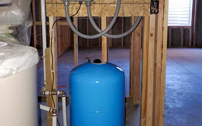 A blue water storage tank is connected to pipes and valves in an unfinished room, part of a residential water pumping system.