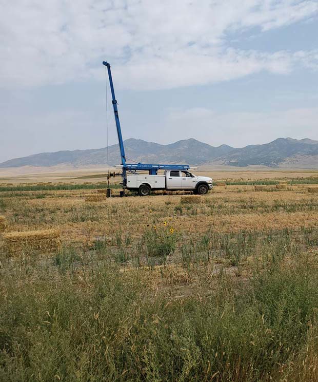 A white truck equipped with a blue crane arm is parked in an open field, likely performing well rehabilitation or maintenance tasks.