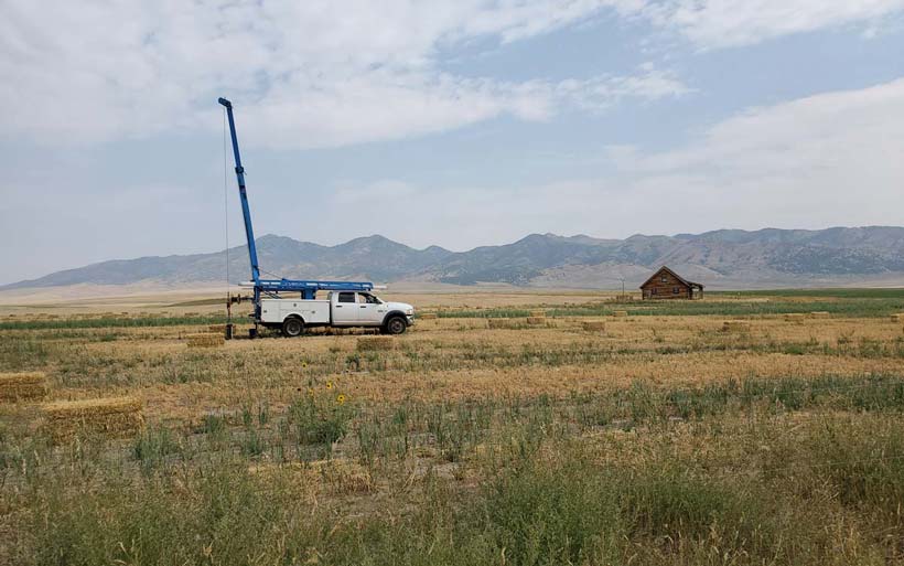 A white truck equipped with a blue crane arm is parked in an open field, likely performing well rehabilitation or maintenance tasks.