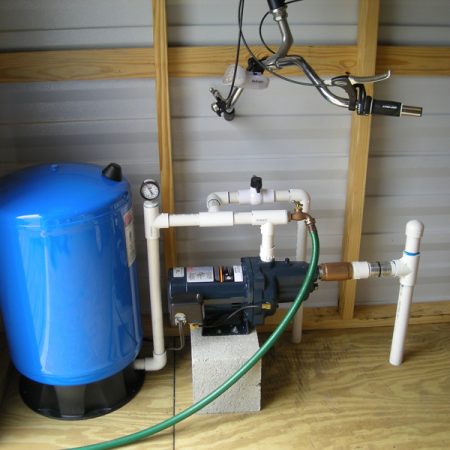 A blue water storage tank is connected to a water pump via pipes and fittings, installed in a room with wooden framing.