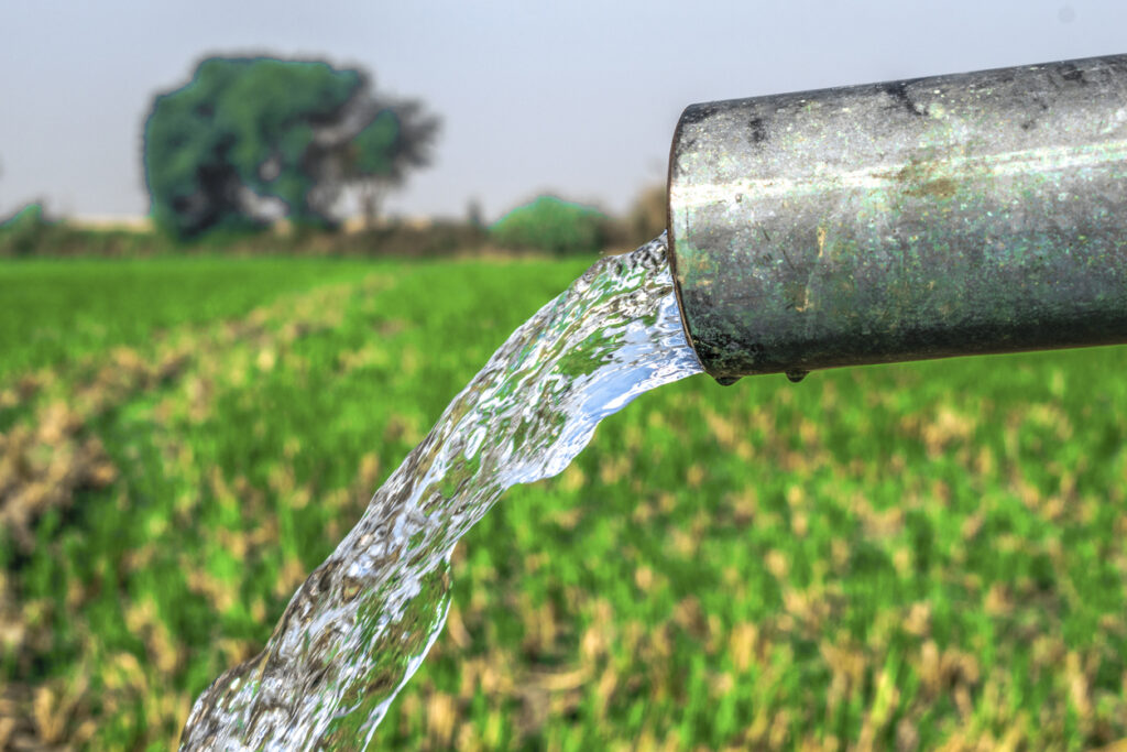 A close-up of water gushing out of a pipe, highlighting its flow into a lush green field with trees in the distance.