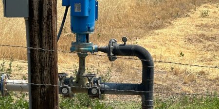 Another view of a blue booster pump mounted on a wooden post near a barbed wire fence, with dry grass and farmland in the background.
