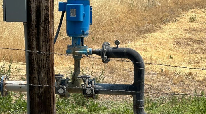 Another view of a blue booster pump mounted on a wooden post near a barbed wire fence, with dry grass and farmland in the background.