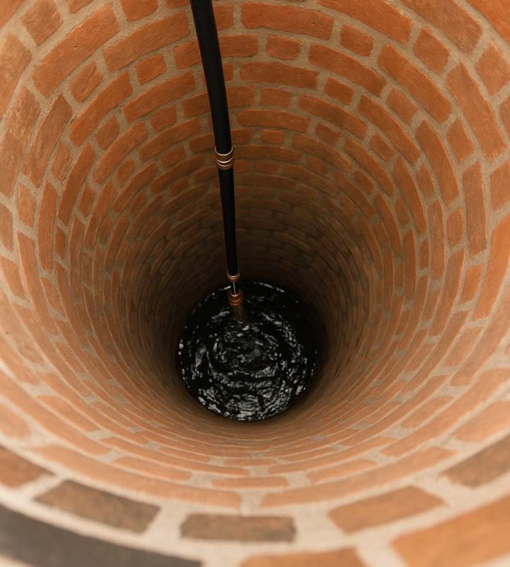 An interior view of a deep brick-lined well with a dark pipe extending downward, showcasing the circular structure and texture of the bricks.