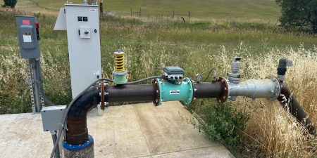A robust water pump system with a control box and various pipes is installed on a concrete slab, surrounded by dry grass and open fields.
