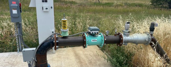 A robust water pump system with a control box and various pipes is installed on a concrete slab, surrounded by dry grass and open fields.