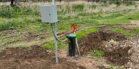 A water pump station with a control box and a green-handled valve is installed outdoors, surrounded by grassy fields and hills.